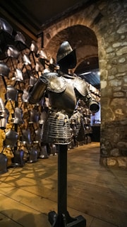 A suit of medieval armor is displayed in a dimly lit room, surrounded by stone walls and wooden flooring. Multiple helmets and breastplates hang in the background, suggesting a historical armory setting.