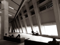 A spacious, modern interior with large angled windows allowing natural light to stream in. Three people are seated on a dark-colored couch facing the windows. Another person is near the windows attending to a child in a stroller. The environment has an industrial feel due to the concrete pillars and ceiling design.