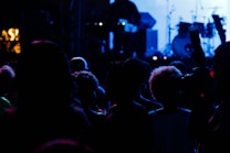 A dark, atmospheric scene featuring a crowd of people silhouetted against a brightly lit stage. The backdrop includes musical instruments and equipment, suggesting a live performance or concert setting. The lighting creates a moody and vibrant ambiance with a mix of blues and purples.