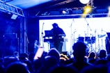 A DJ is performing on stage with a blurred audience in the foreground. The setup includes a laptop and electronic equipment, with drum sets visible in the background. The scene is illuminated with blue and yellow stage lighting, creating an energetic and lively atmosphere.