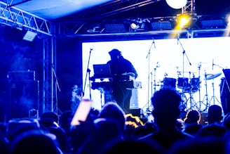 A DJ is performing on stage with a blurred audience in the foreground. The setup includes a laptop and electronic equipment, with drum sets visible in the background. The scene is illuminated with blue and yellow stage lighting, creating an energetic and lively atmosphere.
