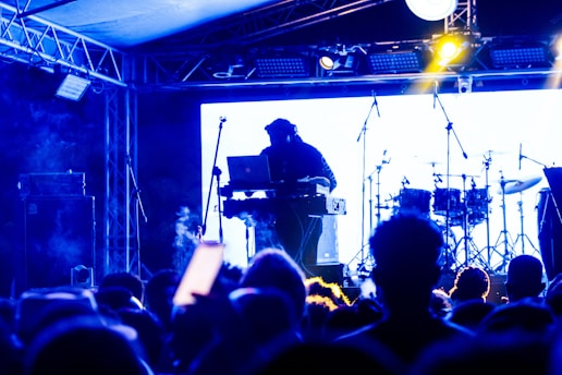 A DJ is performing on stage with a blurred audience in the foreground. The setup includes a laptop and electronic equipment, with drum sets visible in the background. The scene is illuminated with blue and yellow stage lighting, creating an energetic and lively atmosphere.