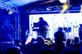 A DJ is performing on stage with a blurred audience in the foreground. The setup includes a laptop and electronic equipment, with drum sets visible in the background. The scene is illuminated with blue and yellow stage lighting, creating an energetic and lively atmosphere.