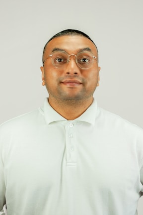 A man with short hair and glasses is wearing a light-colored polo shirt against a plain background.