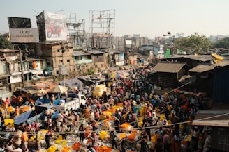 A bustling outdoor market scene with a dense crowd of people engaging in various activities. Vibrant colors of merchandise, particularly bright yellow and orange, are prominent among the stalls. The market is surrounded by makeshift structures and multistory buildings with visible billboards. The atmosphere is lively and crowded, capturing the essence of a busy marketplace.