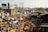 A bustling outdoor market scene with a dense crowd of people engaging in various activities. Vibrant colors of merchandise, particularly bright yellow and orange, are prominent among the stalls. The market is surrounded by makeshift structures and multistory buildings with visible billboards. The atmosphere is lively and crowded, capturing the essence of a busy marketplace.