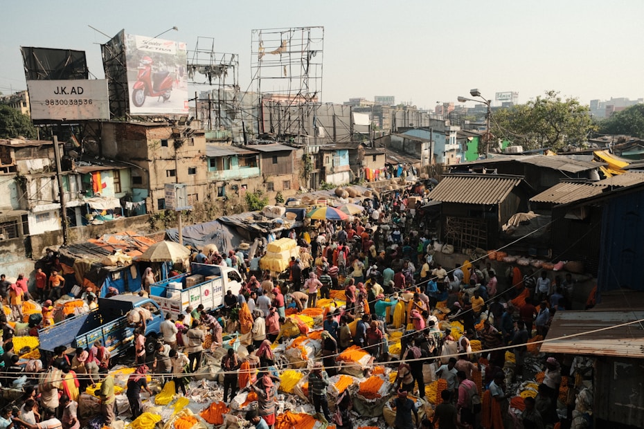 A bustling outdoor market scene with a dense crowd of people engaging in various activities. Vibrant colors of merchandise, particularly bright yellow and orange, are prominent among the stalls. The market is surrounded by makeshift structures and multistory buildings with visible billboards. The atmosphere is lively and crowded, capturing the essence of a busy marketplace.