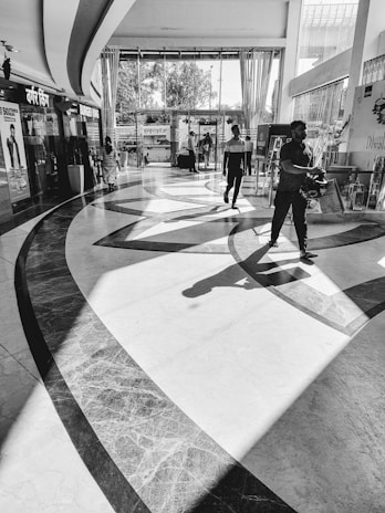 A spacious, modern interior of a shopping area with geometric floor patterns and high ceilings. Natural light pours in through large windows, casting shadows on the floor. There are several people walking in different directions and some shops visible in the background.