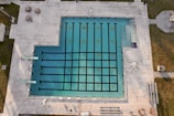 An aerial view of an empty swimming pool with lanes marked by black lines, surrounded by a concrete deck. There are diving boards visible on one side and some poolside furniture, including tables and chairs, placed on the deck area. Grass areas border the concrete deck, and there are some shadows cast across the scene.
