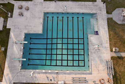An aerial view of an empty swimming pool with lanes marked by black lines, surrounded by a concrete deck. There are diving boards visible on one side and some poolside furniture, including tables and chairs, placed on the deck area. Grass areas border the concrete deck, and there are some shadows cast across the scene.