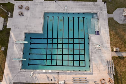 An aerial view of an empty swimming pool with lanes marked by black lines, surrounded by a concrete deck. There are diving boards visible on one side and some poolside furniture, including tables and chairs, placed on the deck area. Grass areas border the concrete deck, and there are some shadows cast across the scene.