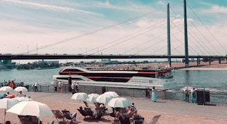 A large passenger boat cruises along a river beneath a modern suspension bridge. Numerous people relax under white umbrellas near the waterfront, enjoying a sunny day. The scene captures a blend of nautical and urban elements, with people casually walking along the promenade.