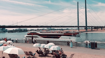 A large passenger boat cruises along a river beneath a modern suspension bridge. Numerous people relax under white umbrellas near the waterfront, enjoying a sunny day. The scene captures a blend of nautical and urban elements, with people casually walking along the promenade.