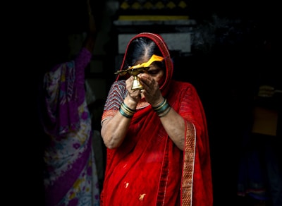 A woman dressed in a vibrant red saree holds a brass lamp with a burning flame, creating a spiritual or ceremonial atmosphere. Her hands are adorned with green bangles, and she is partially obscured by shadows. In the background, another person in a colorful saree is visible, engaged in a similar ritual.