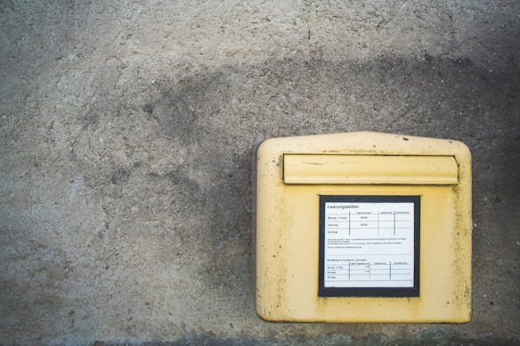 A yellow mailbox is mounted on a weathered concrete wall. The mailbox features a label with collection times and other information written in German.