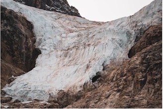 A massive glacier surrounded by rugged rocks, with icy textures and crevices descending from a peak. The ice is layered with a mix of white and pale blue, with some brownish discoloration. The rocky landscape provides a stark contrast to the cold, smooth surface of the glacier.