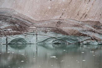 Layers of ice and sediment create a striking pattern on a glacier with a tranquil pool of water in the foreground. The ice exhibits a pale blue hue, while the sediment layers are a mix of brown and gray, forming wavy lines across the glacier surface.