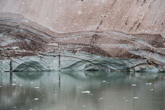 Layers of ice and sediment create a striking pattern on a glacier with a tranquil pool of water in the foreground. The ice exhibits a pale blue hue, while the sediment layers are a mix of brown and gray, forming wavy lines across the glacier surface.