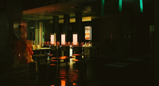 The interior of a fast-food restaurant is depicted in dim lighting, with empty tables and chairs visible. A series of illuminated screens display menu items in the background, casting a warm glow onto the surroundings. Balloons in red and yellow colors are arranged on the left side, suggesting a festive or celebratory occasion.