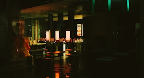 The interior of a fast-food restaurant is depicted in dim lighting, with empty tables and chairs visible. A series of illuminated screens display menu items in the background, casting a warm glow onto the surroundings. Balloons in red and yellow colors are arranged on the left side, suggesting a festive or celebratory occasion.