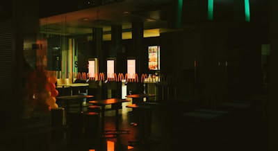 The interior of a fast-food restaurant is depicted in dim lighting, with empty tables and chairs visible. A series of illuminated screens display menu items in the background, casting a warm glow onto the surroundings. Balloons in red and yellow colors are arranged on the left side, suggesting a festive or celebratory occasion.