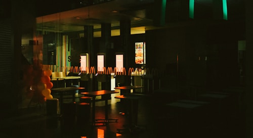The interior of a fast-food restaurant is depicted in dim lighting, with empty tables and chairs visible. A series of illuminated screens display menu items in the background, casting a warm glow onto the surroundings. Balloons in red and yellow colors are arranged on the left side, suggesting a festive or celebratory occasion.