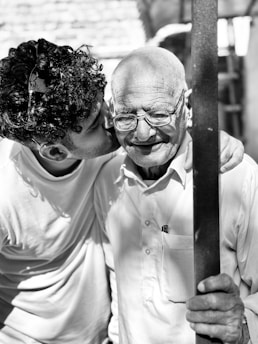 A young man affectionately kisses an elderly man on the cheek. They are close together, with the older man smiling warmly and wearing glasses. Both are dressed in casual attire, and the setting appears to be outdoors with natural lighting.