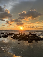 the sun is setting over the ocean with rocks in the foreground