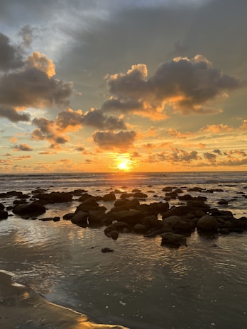 the sun is setting over the ocean with rocks in the foreground