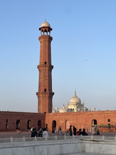 A tall, reddish-brown minaret stands prominently under a clear blue sky. In the background, a white domed structure with intricate spires is visible, suggesting an architectural blend of Islamic and Mughal influences. People are scattered across a wide open area, some standing and others walking, adding a sense of scale to the scene.