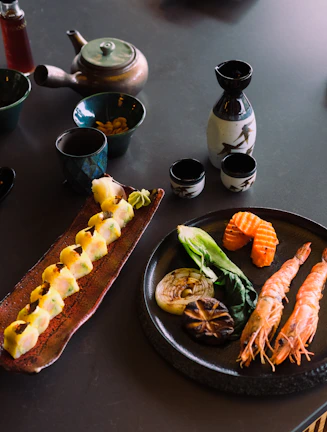 A variety of Japanese dishes on a table, including a plate with fresh sushi rolls, grilled vegetables, and prawns. There is also a teapot and cups alongside a bottle of sake with small sake cups.