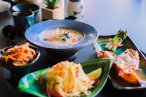 A table displays an assortment of dishes, including a creamy soup in a blue bowl adorned with herbs and spices, a side dish with asparagus and vegetables, and a vibrant salad garnished with lime. The setting suggests a fine dining or sophisticated culinary experience.