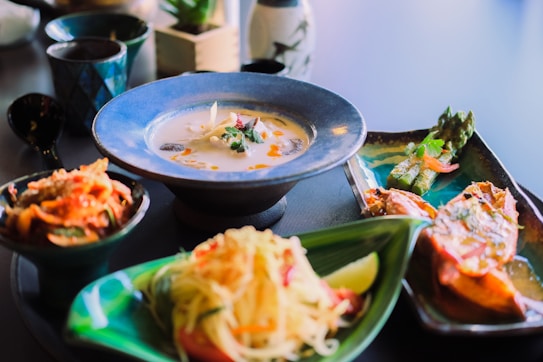 A table displays an assortment of dishes, including a creamy soup in a blue bowl adorned with herbs and spices, a side dish with asparagus and vegetables, and a vibrant salad garnished with lime. The setting suggests a fine dining or sophisticated culinary experience.