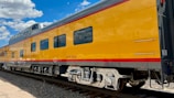 A bright yellow train car with windows reflecting blue skies and clouds is seen on train tracks. The train has a red stripe along the bottom and top, with the words 'Colorado Eagle' written on the side.