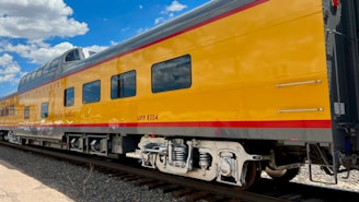 A bright yellow train car with windows reflecting blue skies and clouds is seen on train tracks. The train has a red stripe along the bottom and top, with the words 'Colorado Eagle' written on the side.