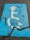 A weathered blue parking space marking on asphalt depicting a person in a wheelchair with a graduation cap and holding a diploma. The paint is cracked and faded, with a prominent black crack running across it.