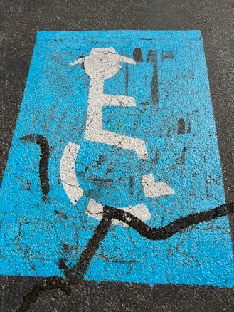 A weathered blue parking space marking on asphalt depicting a person in a wheelchair with a graduation cap and holding a diploma. The paint is cracked and faded, with a prominent black crack running across it.