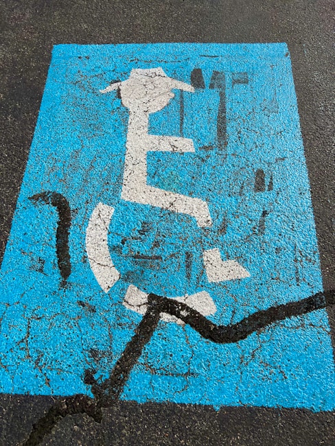 A weathered blue parking space marking on asphalt depicting a person in a wheelchair with a graduation cap and holding a diploma. The paint is cracked and faded, with a prominent black crack running across it.