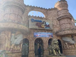 A theme park entrance styled like an ancient castle with decorative towers and arches. The facade features sculptures of mythical creatures, including winged figures and a large lion. The signage reads 'Dream World Water Park.' Garlands and lights hang from the structure.