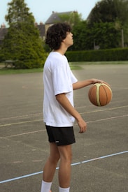 A person is standing on an outdoor basketball court, casually holding a basketball in one hand. They are wearing a white t-shirt, black shorts, and white socks. The background features green trees and a couple of residential rooftops, indicating a suburban setting.