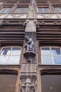 An intricately carved wooden facade features a statue of a person in historical attire. The architecture includes detailed window frames and decorative elements, creating a classic and artistic appearance. The craftsmanship is ornate, reflecting a style from a bygone era.
