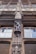 An intricately carved wooden facade features a statue of a person in historical attire. The architecture includes detailed window frames and decorative elements, creating a classic and artistic appearance. The craftsmanship is ornate, reflecting a style from a bygone era.