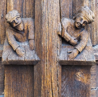 Two wooden carved figures are depicted on a rustic textured surface. The figures have detailed facial expressions and are positioned as if leaning or emerging from the wood. The wood displays rich, natural grains and a weathered appearance.