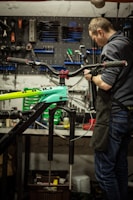 Inside a workshop, a person is focused on repairing or assembling a bicycle. Various tools are organized on a pegboard in the background, along with other bicycle parts at a workbench. The bicycle frame is prominently visible in the foreground.
