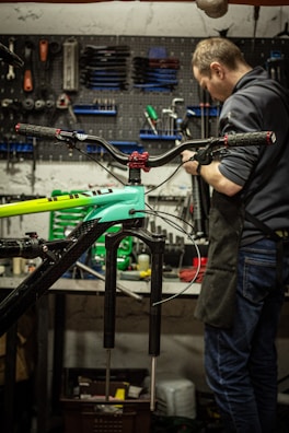 Inside a workshop, a person is focused on repairing or assembling a bicycle. Various tools are organized on a pegboard in the background, along with other bicycle parts at a workbench. The bicycle frame is prominently visible in the foreground.