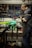 Inside a workshop, a person is focused on repairing or assembling a bicycle. Various tools are organized on a pegboard in the background, along with other bicycle parts at a workbench. The bicycle frame is prominently visible in the foreground.