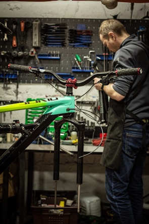 Inside a workshop, a person is focused on repairing or assembling a bicycle. Various tools are organized on a pegboard in the background, along with other bicycle parts at a workbench. The bicycle frame is prominently visible in the foreground.