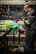 Inside a workshop, a person is focused on repairing or assembling a bicycle. Various tools are organized on a pegboard in the background, along with other bicycle parts at a workbench. The bicycle frame is prominently visible in the foreground.