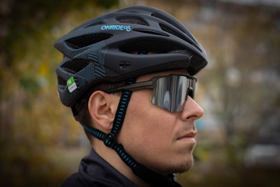 A person wearing a black cycling helmet paired with dark, reflective sunglasses. The helmet has blue accents and a green sticker indicating size. The background is softly blurred, suggesting an outdoor setting.