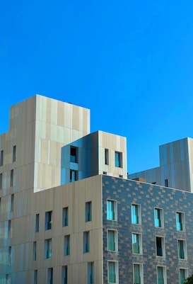 A modern building with a geometric design featuring a mix of rectangular and square shapes. The facade is composed of light beige and dark textured panels, interspersed with numerous windows that reflect the bright blue sky.